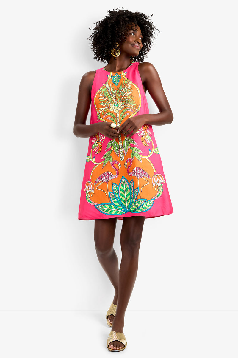 A woman smiles against a white background, wearing the Loveage Dress—a sleeveless, vibrant pink piece with colorful tropical prints and flamingos. She accessorizes with gold earrings and beige sandals, touching her curly hair.