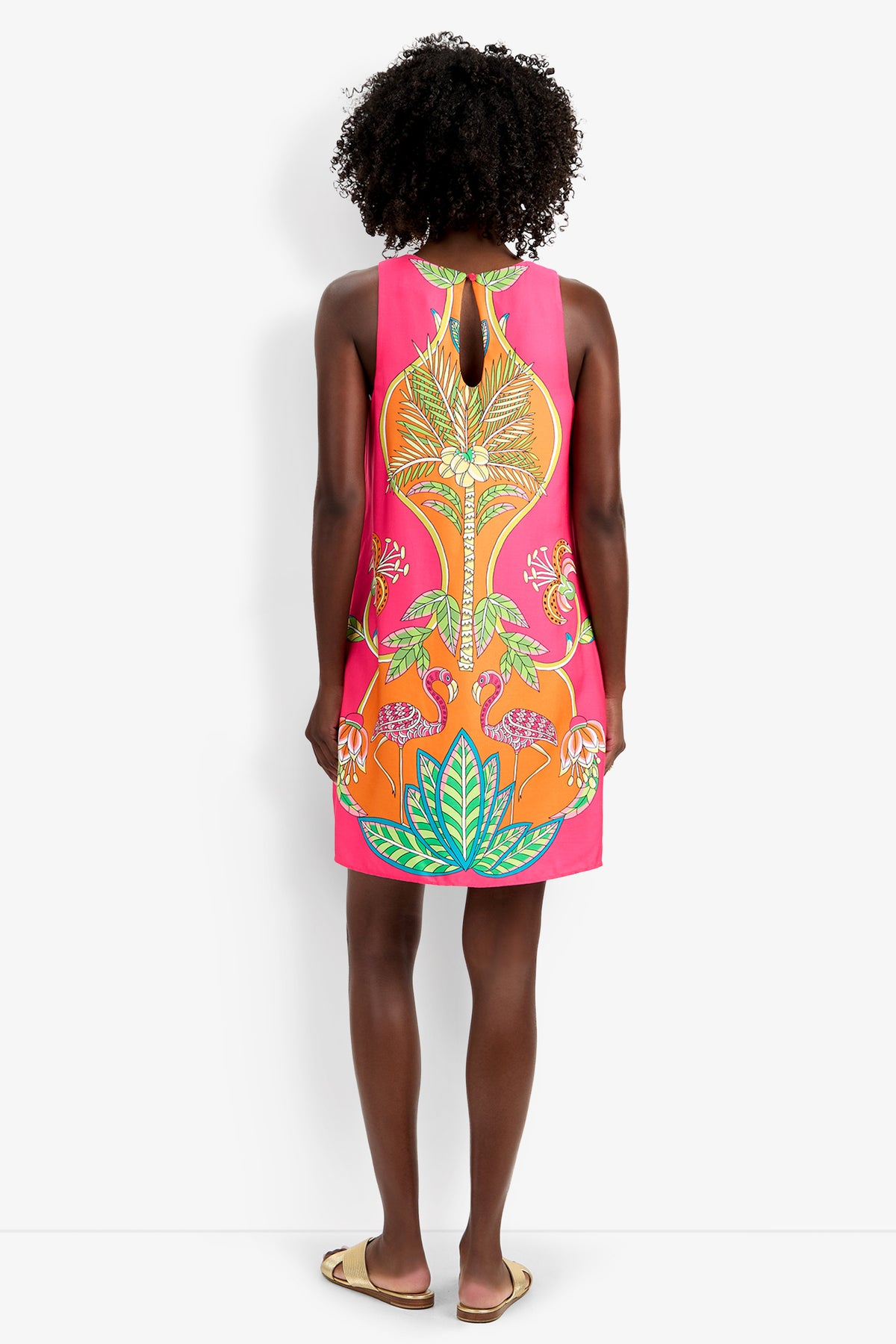 A woman stands facing away, wearing the sleeveless Loveage Dress with a vibrant tropical leaf and flamingo design, paired with flat sandals.