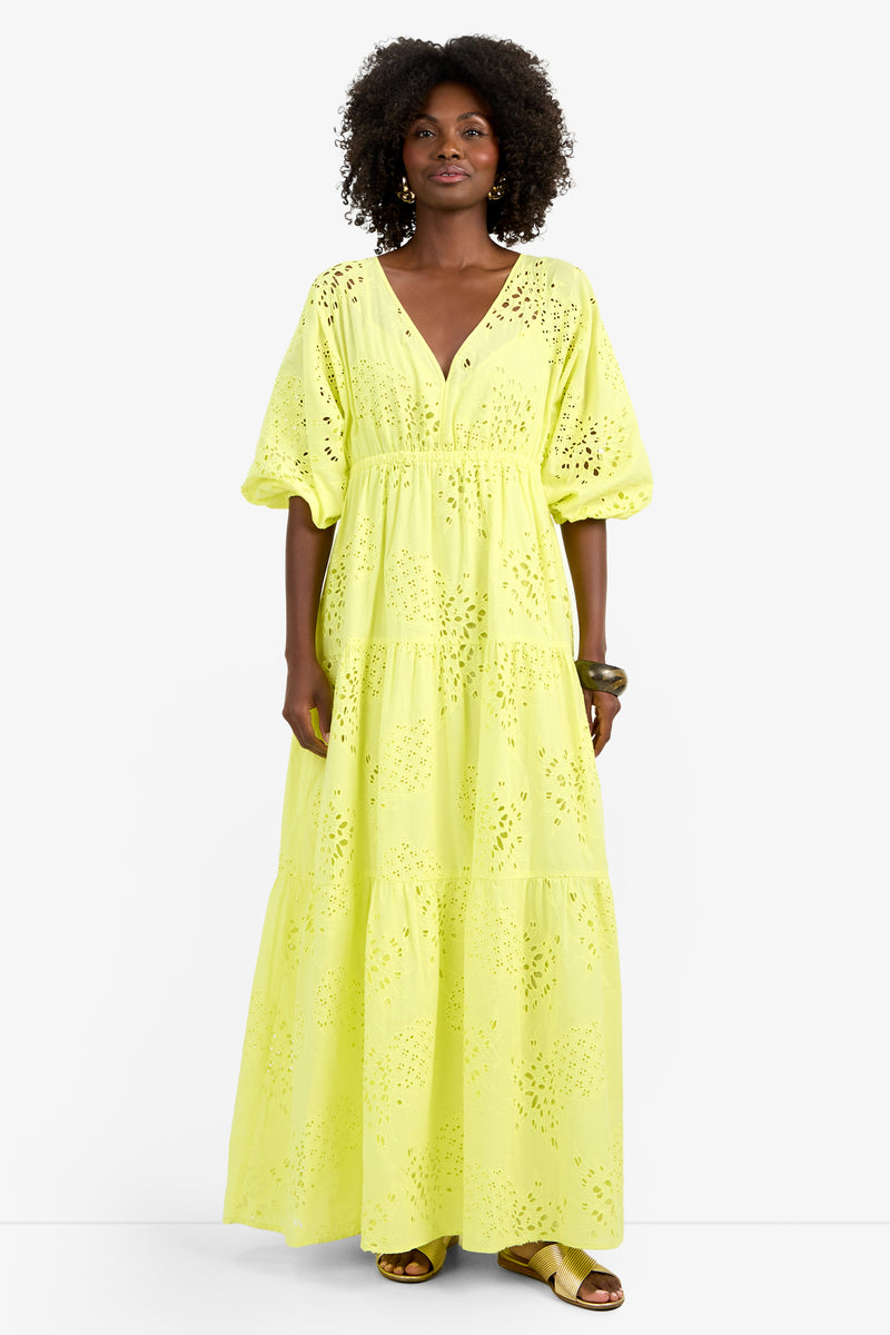 A woman with curly hair wears the ARCO IRIS DRESS, an ankle-length bright yellow 100% cotton eyelet piece with puffed sleeves, V-neckline, and tiered skirt. She pairs it with sandals and a chunky bracelet against a plain white background.