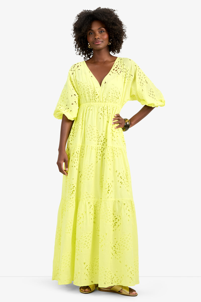 A woman stands against a white background wearing the ARCO IRIS DRESS, a long yellow 100% cotton eyelet dress with puffy sleeves and a V-neckline, paired with earrings, a bracelet, and sandals.