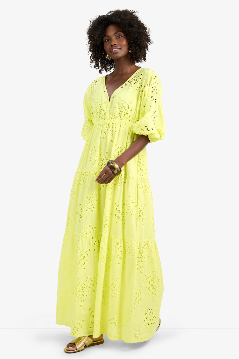 A woman with curly hair wears the ARCO IRIS DRESS—a long, bright yellow 100% cotton eyelet dress with puffed three-quarter sleeves and a V-neckline—paired with gold sandals and bangles, standing against a plain white background.