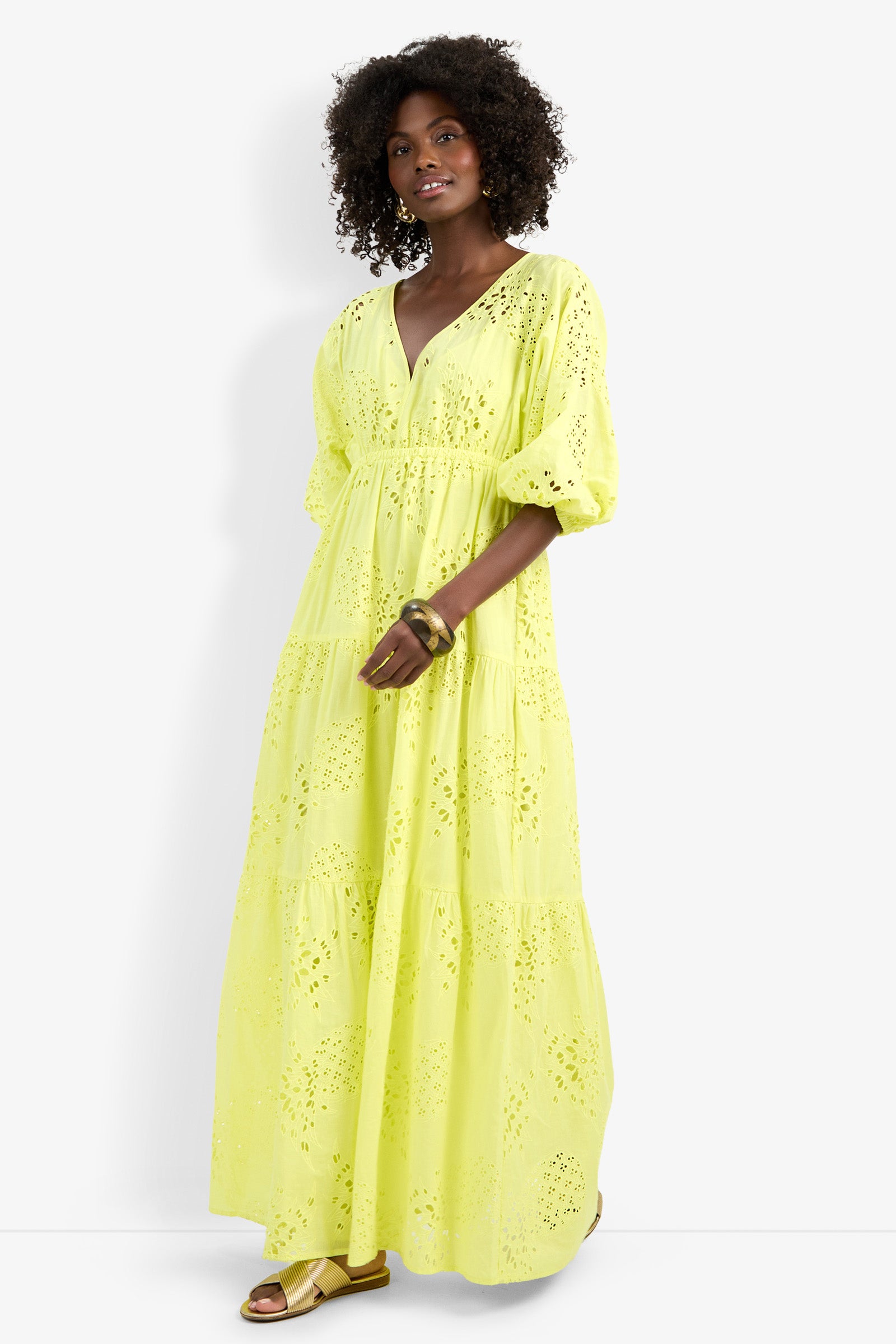 A woman with curly hair wears the Arco Iris Dress—a long, bright yellow eyelet dress with puffed sleeves and a V-neck—paired with gold sandals and bangles, standing against a plain white background.