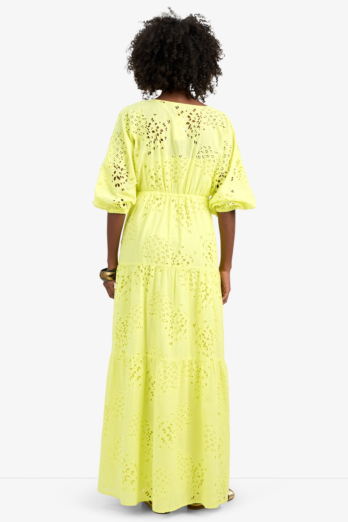 A person with curly hair, facing away, wears the ARCO IRIS DRESS—a long, tiered, light yellow 100% cotton eyelet dress with elbow-length sleeves, cut-out patterns, and a relaxed fit.