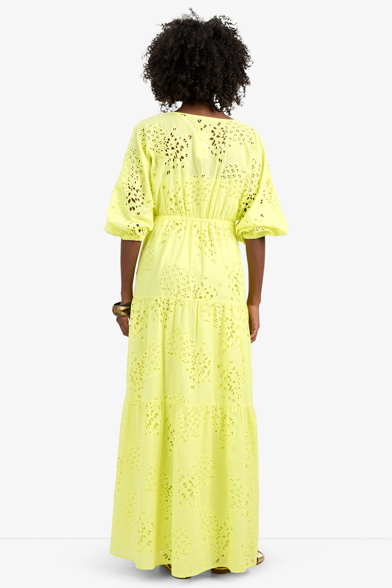 A person with curly hair, facing away, wears the ARCO IRIS DRESS—a long, tiered, light yellow 100% cotton eyelet dress with elbow-length sleeves, cut-out patterns, and a relaxed fit.