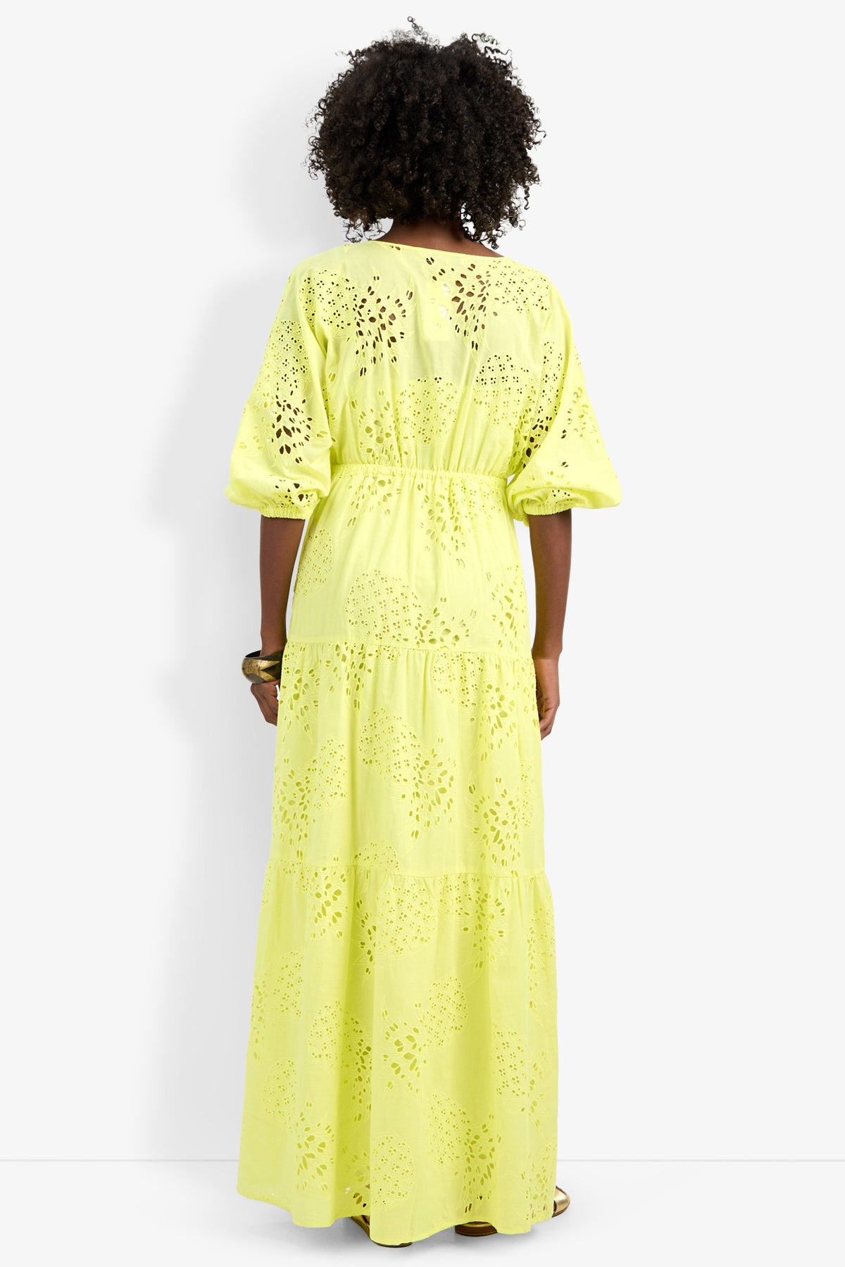 A person with curly hair stands with their back to the camera in a long, yellow Arco Iris Dress with puffed sleeves, set against a white background.