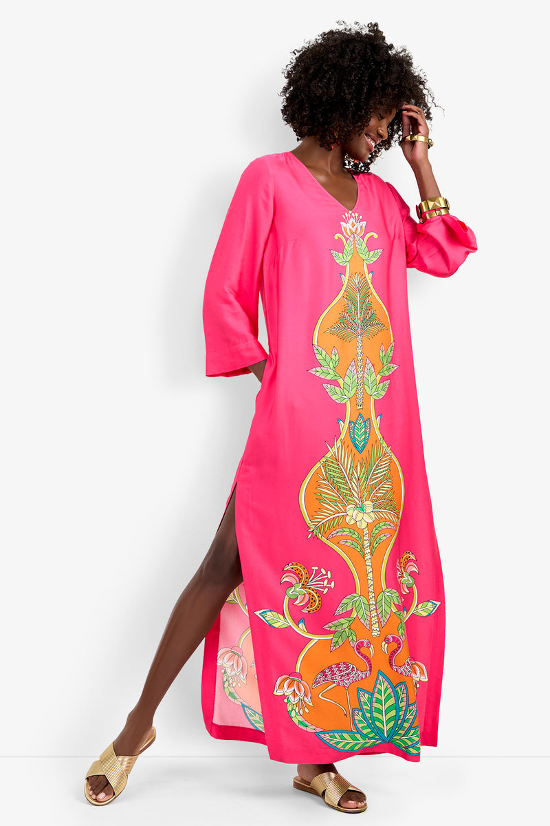 A woman smiles sideways in the Lanessa Dress, a bright pink maxi with bold orange and green botanicals and a high side slit. She accessorizes with gold bracelets and sandals.