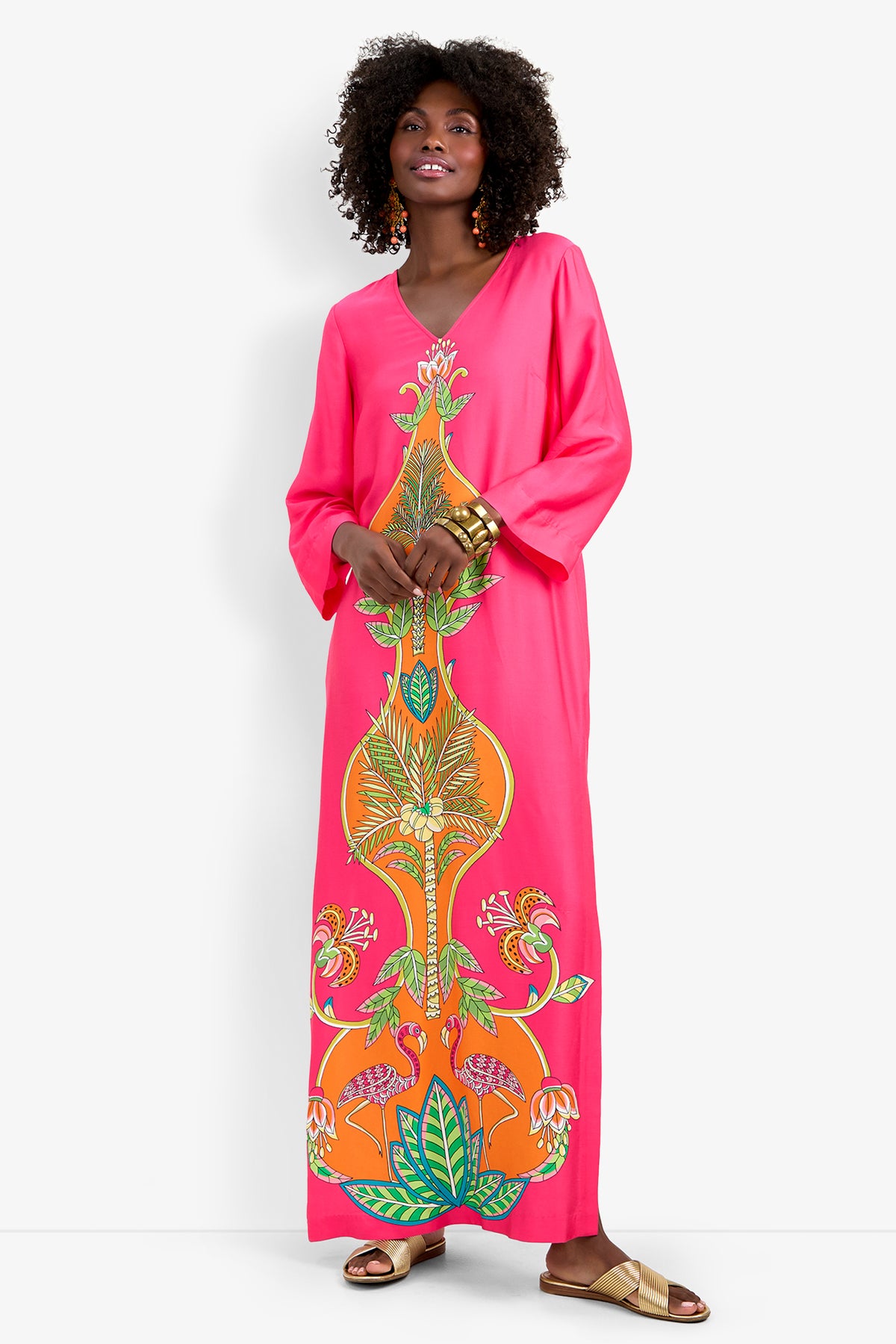 A woman wearing the Lanessa Dress, a long bright pink dress with colorful floral and leaf patterns, gold bracelets, and gold sandals stands smiling against a plain white background with her hands at her sides.