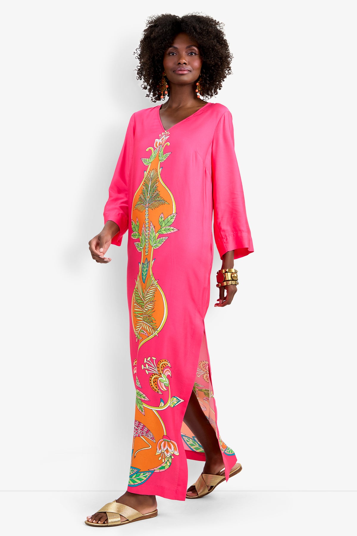 A woman walks confidently in the Lanessa Dress, a bright pink long-sleeve maxi with a colorful tropical floral pattern, accessorized with gold bangles and sandals against a plain white background.