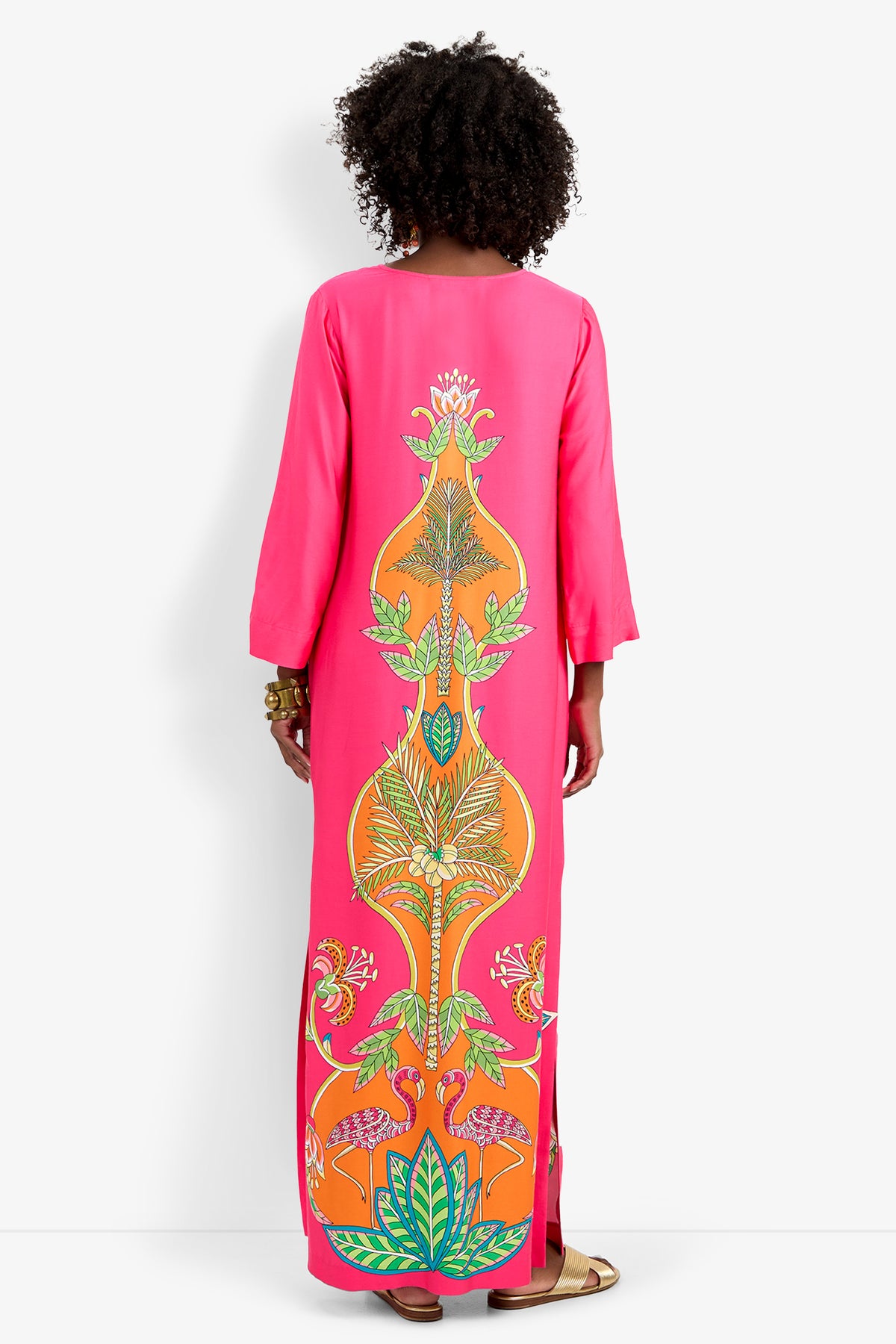A woman with curly hair, sandals, and bracelets is seen from behind wearing the Lanessa Dress—a long, bright pink piece featuring a vivid tropical floral pattern in orange, green, and yellow on the back.