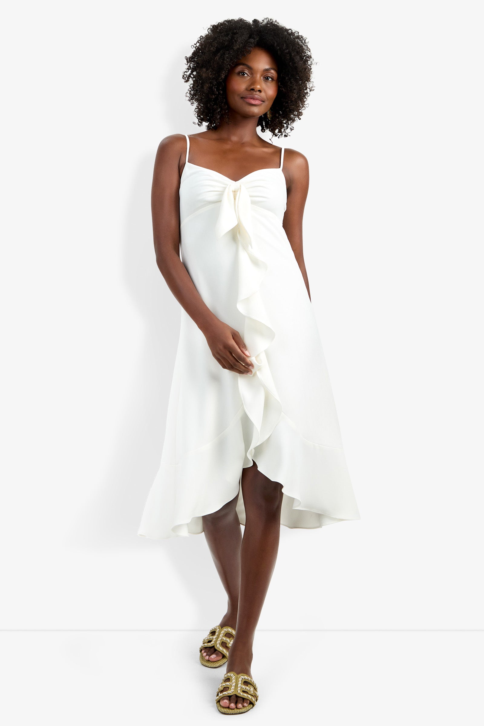 A woman stands against a plain white background, wearing the Lilla Dress—a sleeveless white midi dress with ruffle detail down the front—and gold sandals.