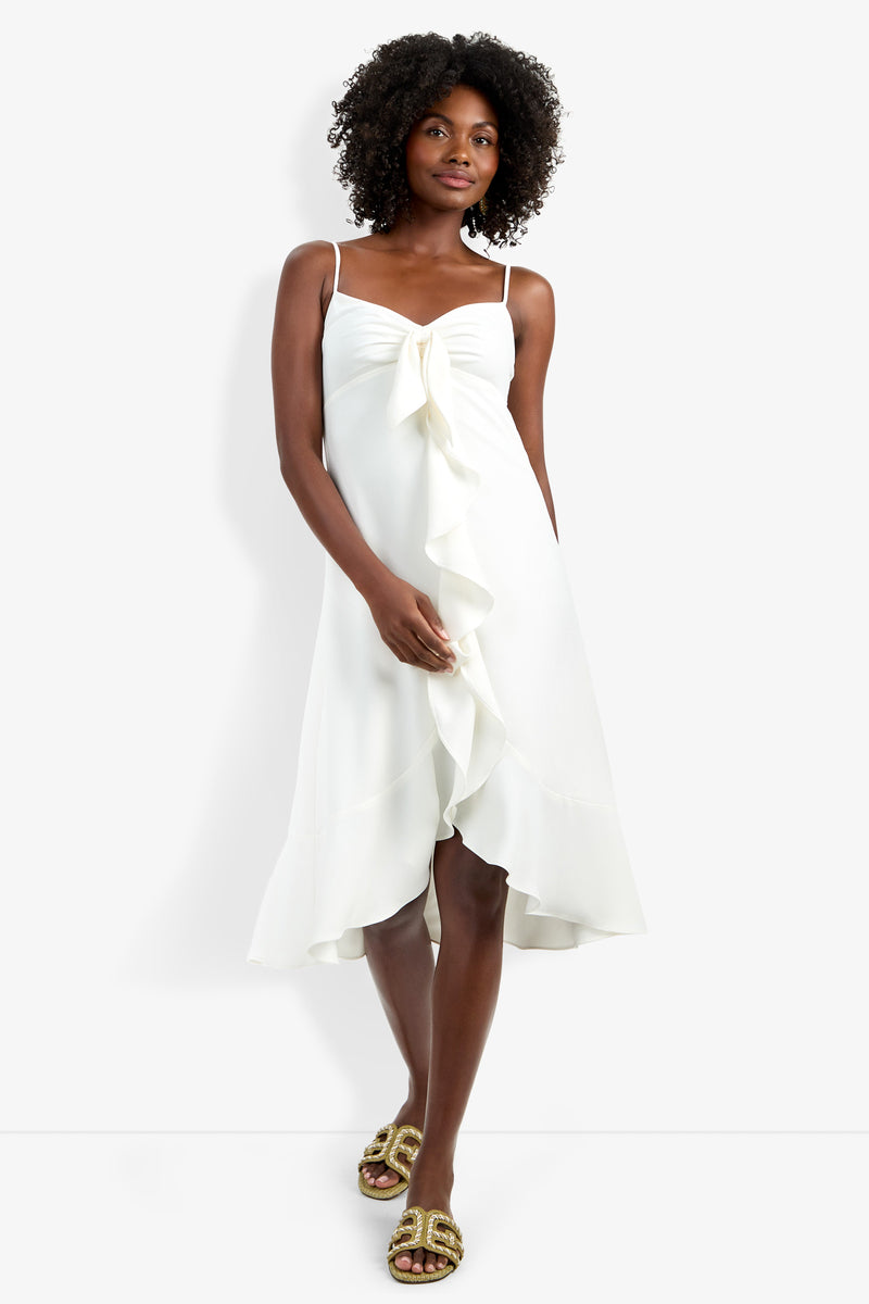 A woman with curly hair wears the Lilla Dress, a sleeveless, knee-length white dress featuring a front ruffle detail and bow at the bust, paired with gold slide sandals against a plain background.
