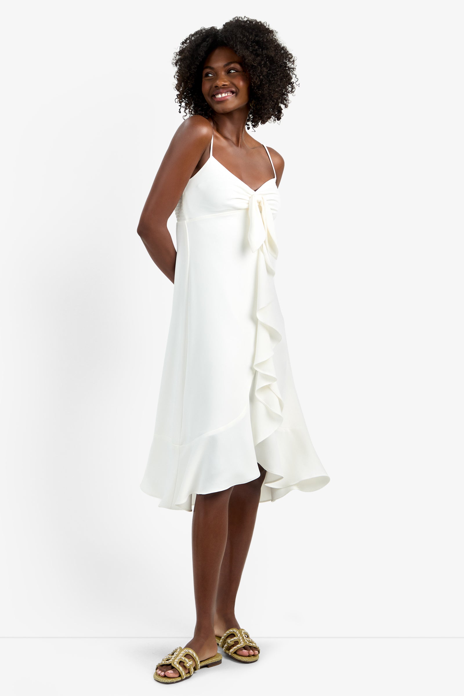 A woman with curly hair smiles in the LILLA DRESS—a white, knee-length, sleeveless dress with ruffle detail and spaghetti straps, made from 100% polyester. She pairs it with gold sandals against a plain white background.