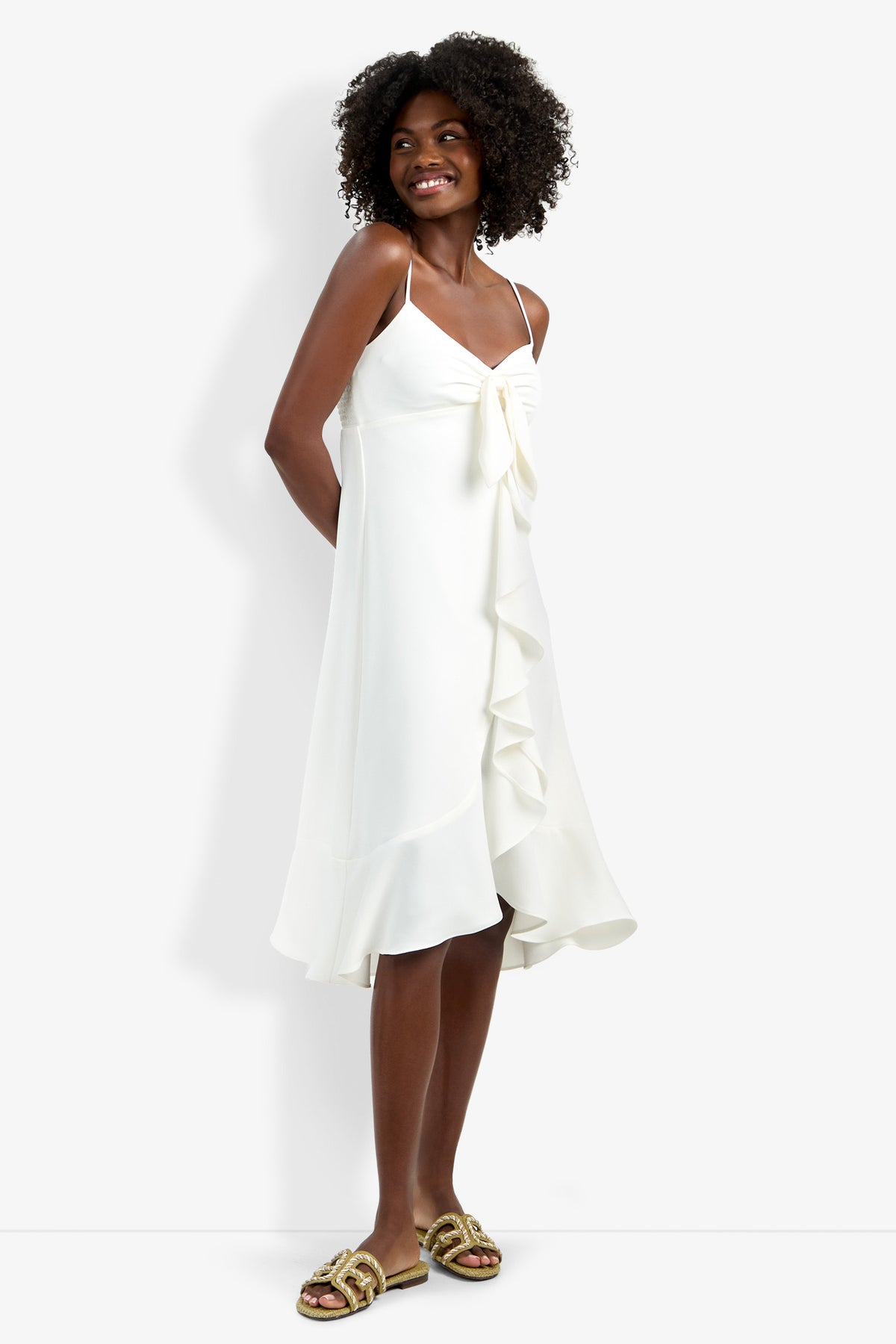 A woman stands smiling in the Lilla Dress—a white, sleeveless, knee-length dress with thin straps and ruffle details—her arms relaxed and feet in gold sandals.