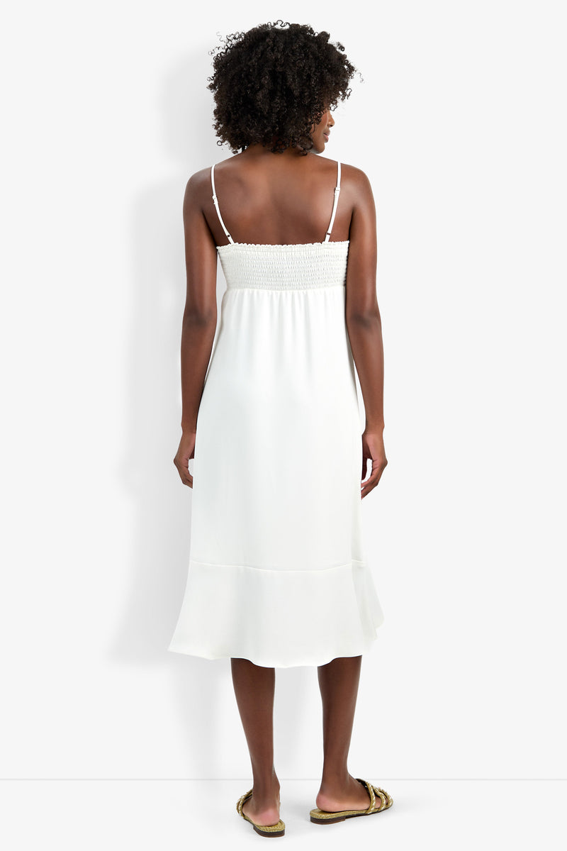 A woman with curly hair faces away, wearing the Lilla Dress—a white, sleeveless, midi-length dress with thin straps and a ruffled hem—paired with flat sandals against a plain white background.