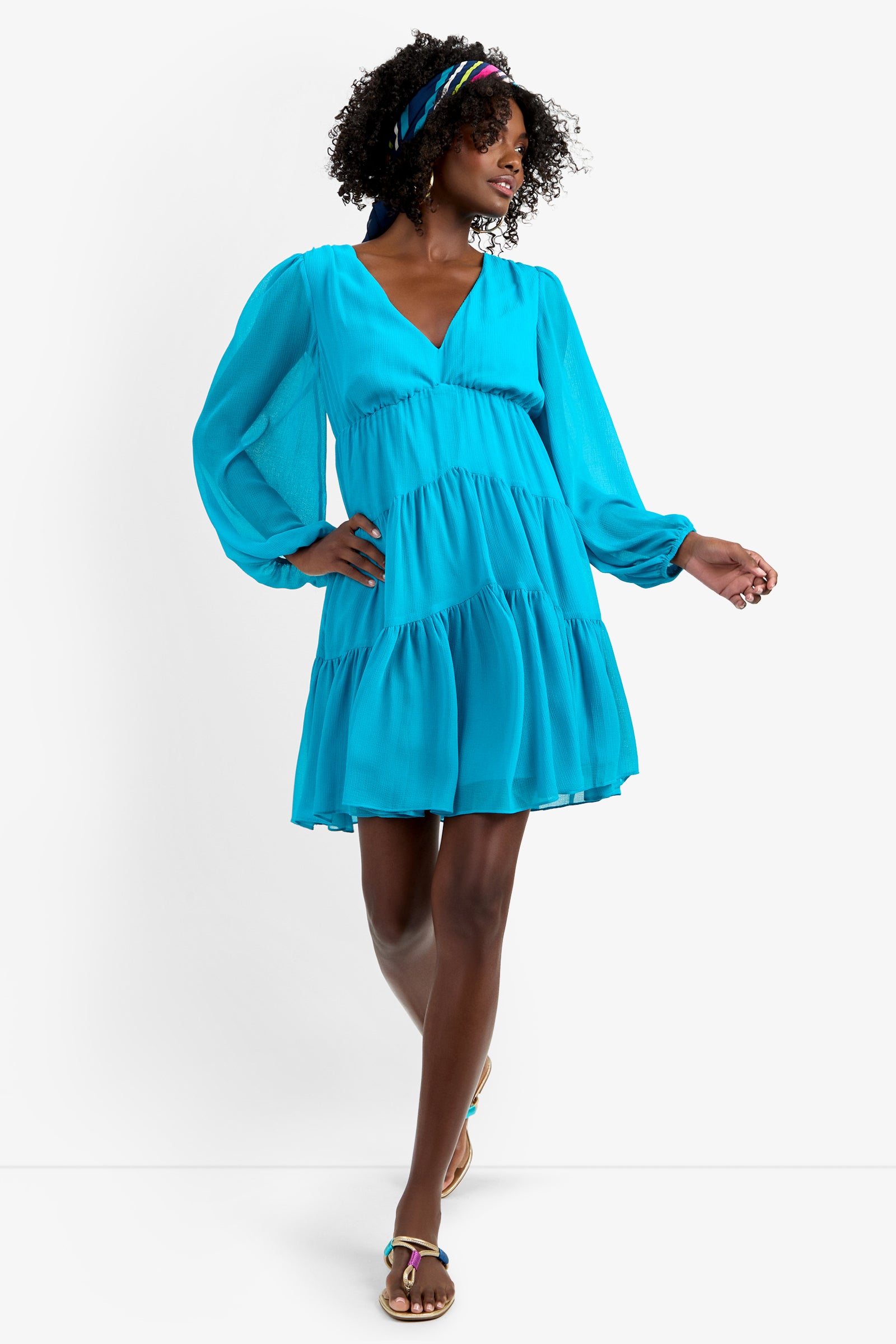 A woman poses confidently in the MAKE MERRY DRESS, a bright blue, long-sleeve, tiered mini dress made of 100% polyester. She pairs it with sandals and a colorful headband, smiling with one hand on her hip against a plain white background.