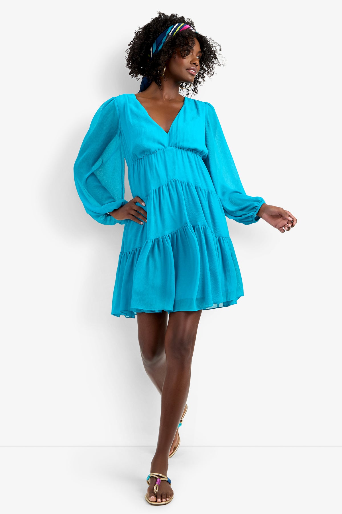 A woman with curly hair and a colorful headband smiles while standing with one leg crossed, wearing the Make Merry Dress—a bright blue, long-sleeve, tiered mini dress—and sandals.