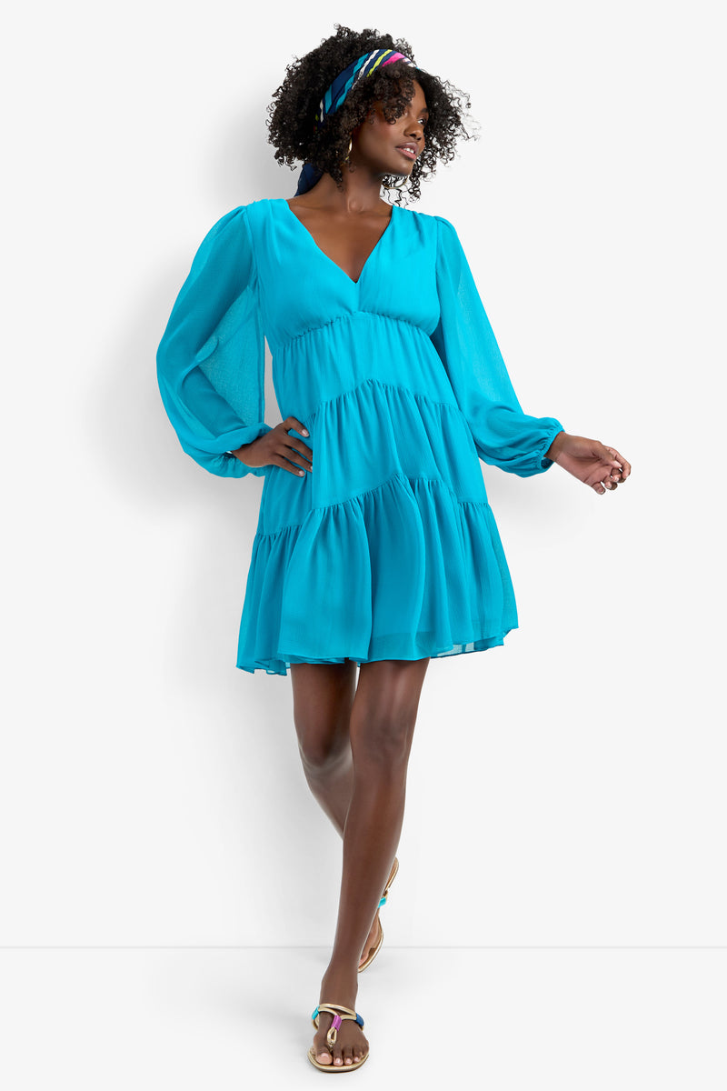 A woman with curly hair and a colorful headband poses against a white background, wearing the Make Merry Dress— a bright blue, long-sleeve, tiered mini dress—paired with sandals and one hand raised.