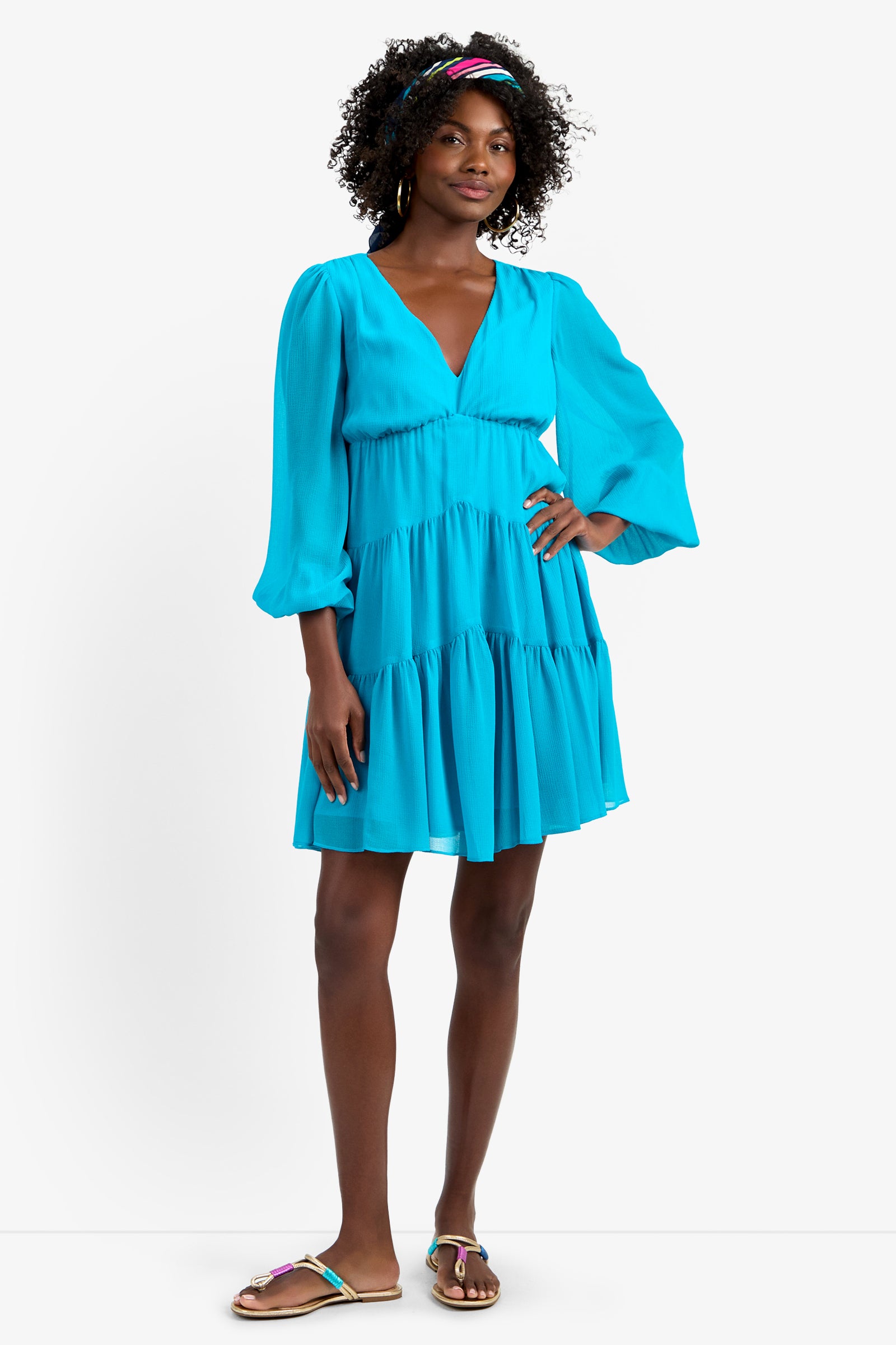 A woman in the MAKE MERRY DRESS—a bright blue, long-sleeve, V-neck tiered dress in soft polyester—stands with one hand on her hip, curly hair held back by a colorful headband, and wears sandals.