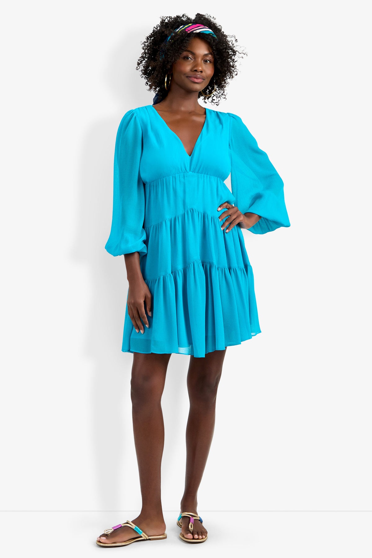 A woman with curly hair in a colorful headband smiles, wearing the Make Merry Dress—a bright blue, long-sleeve, tiered V-neck dress with puff sleeves—and sandals against a plain white background.