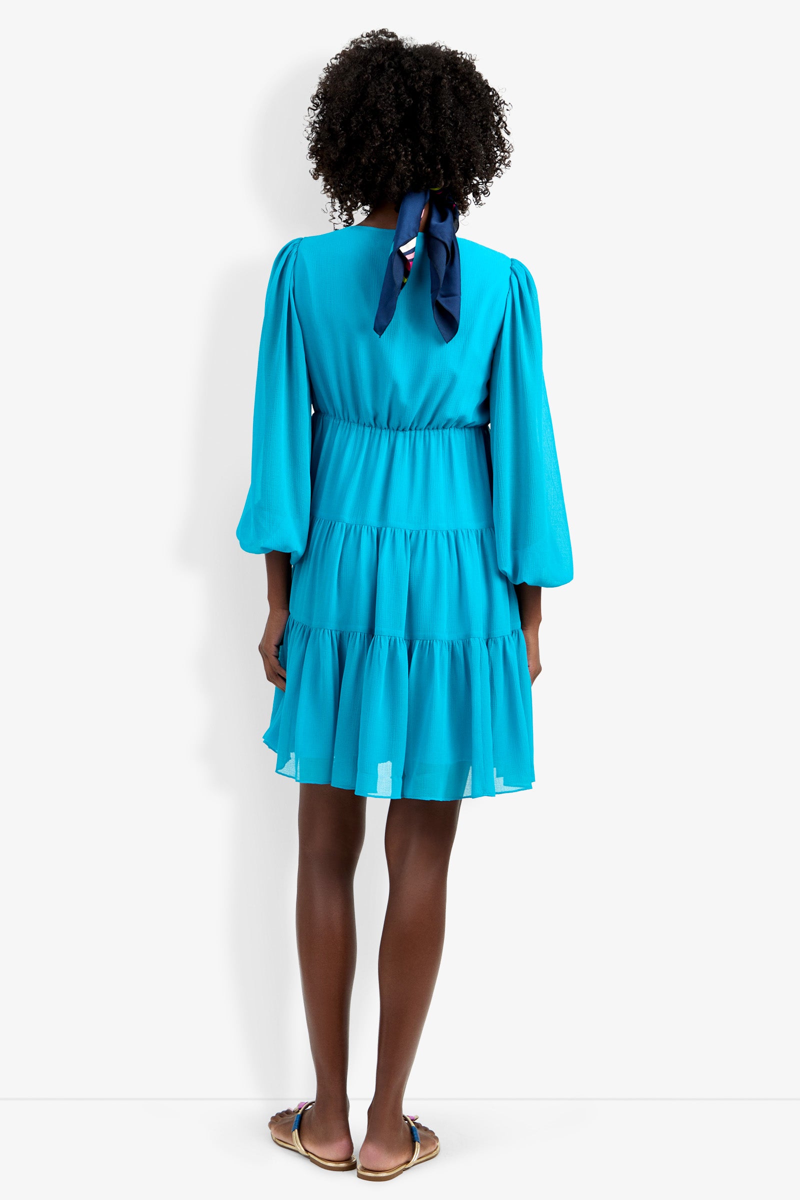A person faces away, wearing the Make Merry Dress in bright blue with long sleeves and tiers, paired with flat sandals. Their curly hair is tied back with a navy ribbon against a plain white background.