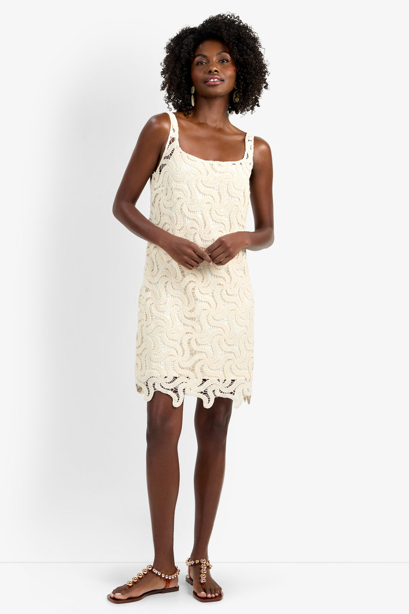 A woman wearing the ASPIRE DRESS— a sleeveless, cream lace cotton dress with wavy patterns and a scalloped hem—stands smiling, her curly hair framing her face. She pairs it with flat brown sandals and clasps her hands gently.