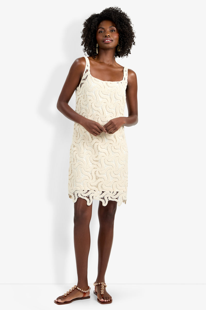 A woman with curly hair wears the sleeveless, cream Aspire Dress featuring a wavy crochet pattern and scalloped hem, paired with beaded flat sandals, standing against a white background.