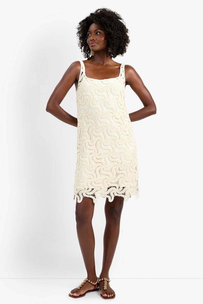 A woman with curly hair wears the ASPIRE DRESS—a sleeveless, cream crochet dress in 100% cotton with a wavy pattern and scalloped hem—paired with embellished sandals, standing against a white background.