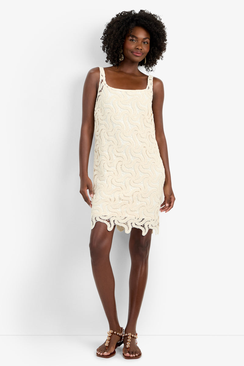 A woman with curly hair wears the ASPIRE DRESS, a cream, sleeveless crochet mini dress in 100% cotton featuring a wavy pattern and scalloped hem, paired with studded flat sandals against a white background.