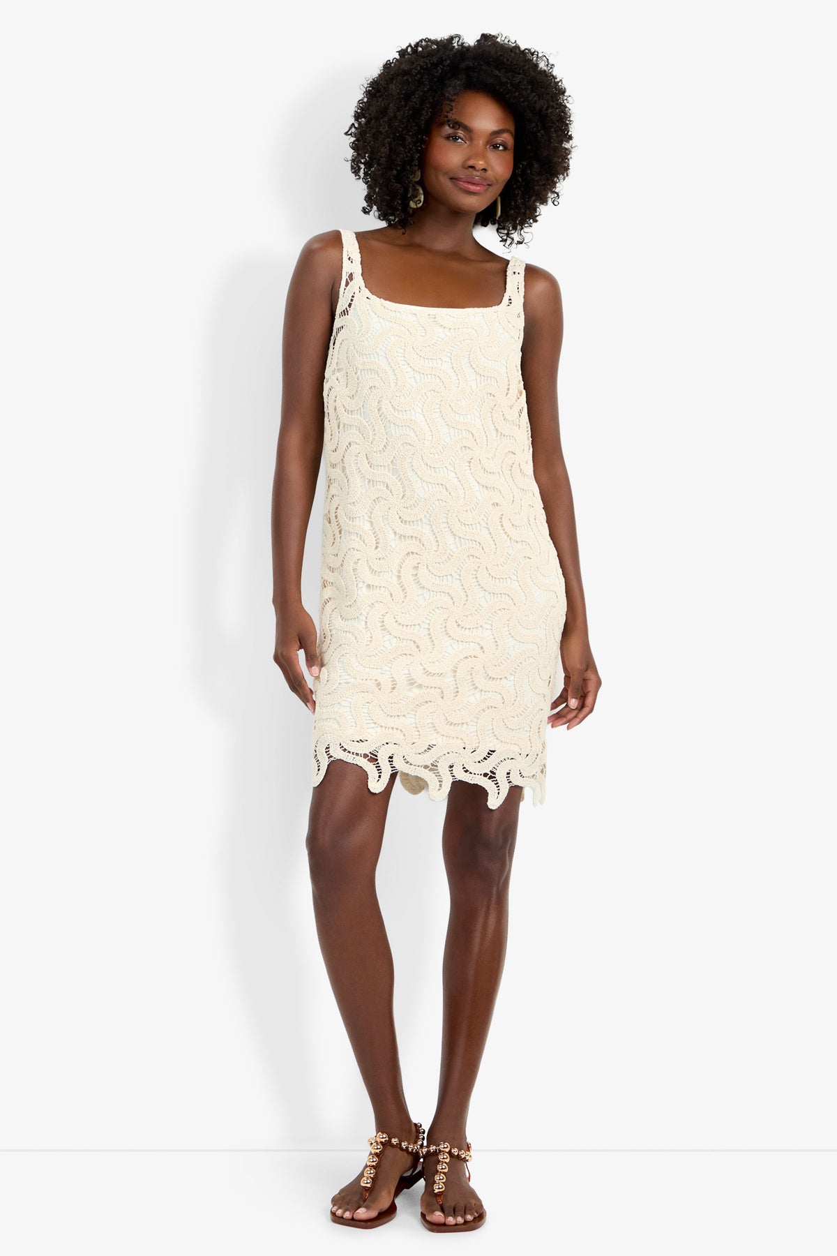 A woman with curly hair wears the Aspire Dress—a sleeveless, cream crochet dress with a wavy pattern and scalloped hem—paired with studded flat sandals, posing against a white background.