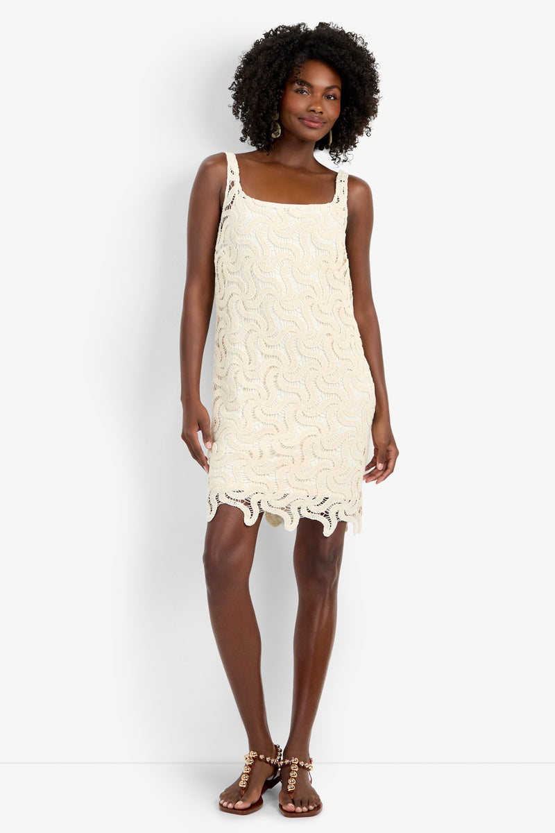A woman with curly hair wears the sleeveless, cream Aspire Dress featuring a wavy crochet pattern and scalloped hem, paired with strappy flat sandals. She stands against a white background, smiling gently.