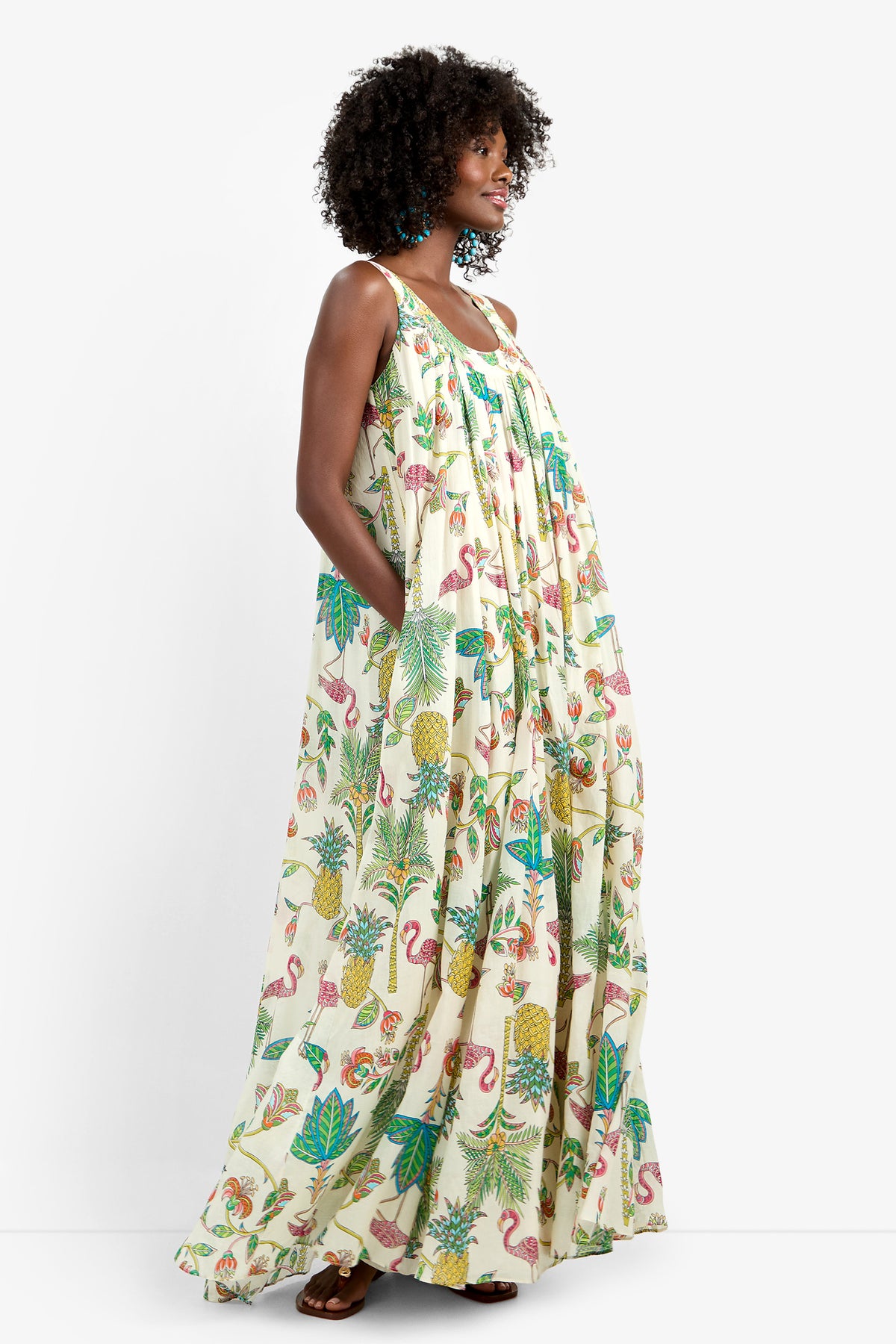 A woman stands sideways with her hand in her pocket, wearing the JIANNA MAXI DRESS—a long 100% cotton dress featuring a colorful tropical print with pineapples, flamingos, and leaves—against a plain white background.