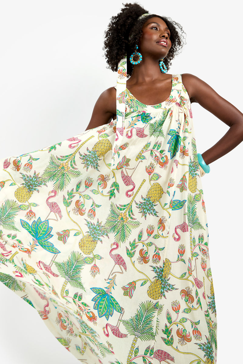 A woman smiles and poses in the JIANNA MAXI DRESS, a sleeveless, flowing 100% cotton dress with vibrant tropical prints of pineapples, flamingos, and leaves. She accessorizes with turquoise earrings and a matching headband, hand on her hip.