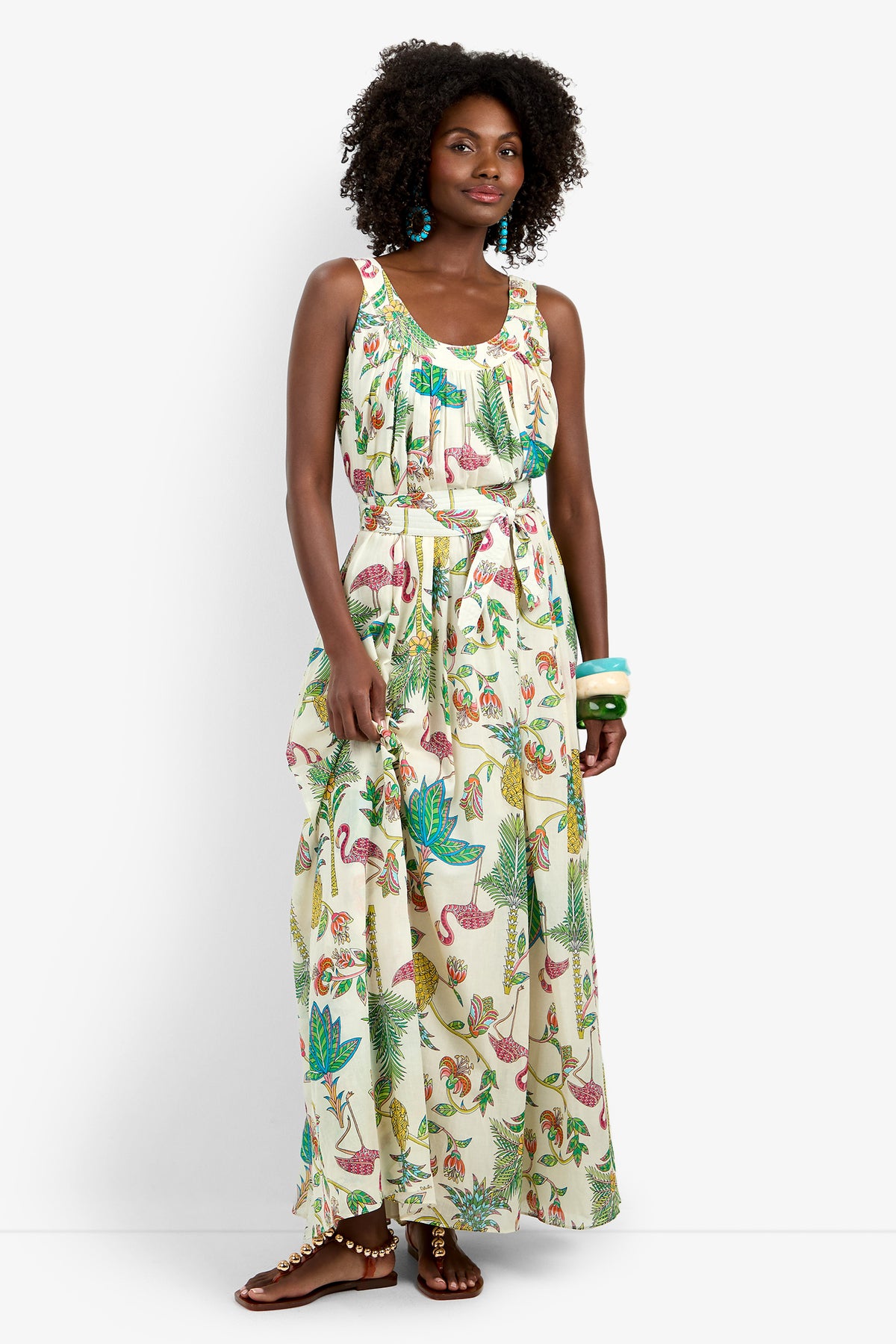 A woman wears the JIANNA MAXI DRESS, a sleeveless, ankle-length 100% cotton dress with a tropical flamingo and green leaf print. She pairs it with turquoise earrings, chunky bracelets, and brown sandals.