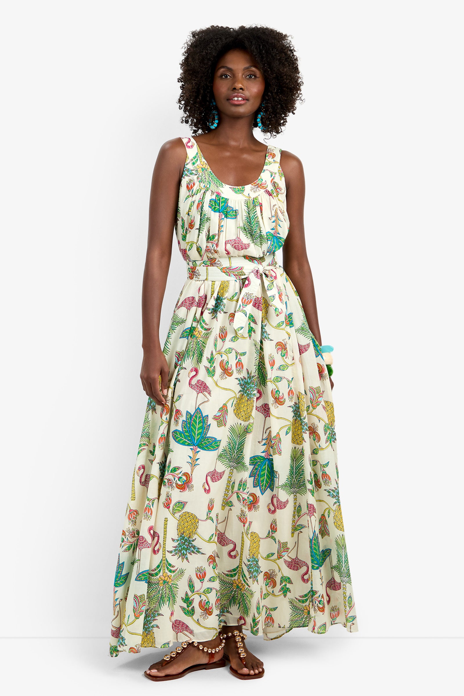 A woman wears the JIANNA MAXI DRESS in MULTI, a sleeveless, floor-length 100% cotton gown with a vibrant tropical print of leaves, flamingos, pineapples, and parrots. She accessorizes with turquoise earrings, a bracelet, and brown sandals.