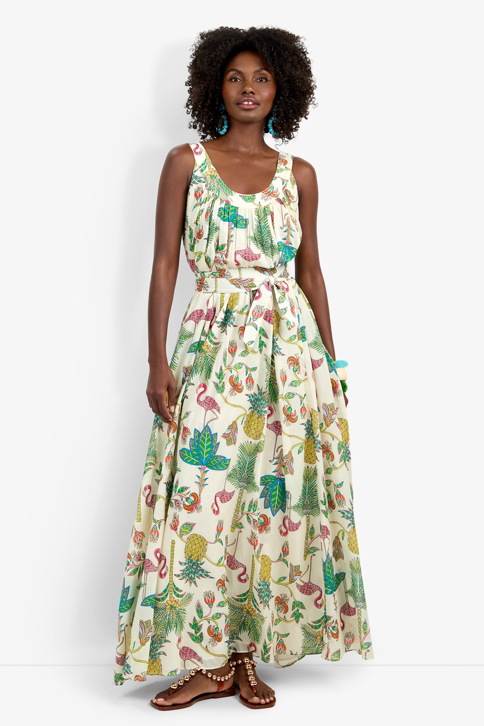 A woman wears the Jianna Maxi Dress, a sleeveless, floor-length dress with a tropical print of flamingos, palm leaves, and pineapples. She has curly hair, turquoise earrings, and brown sandals.