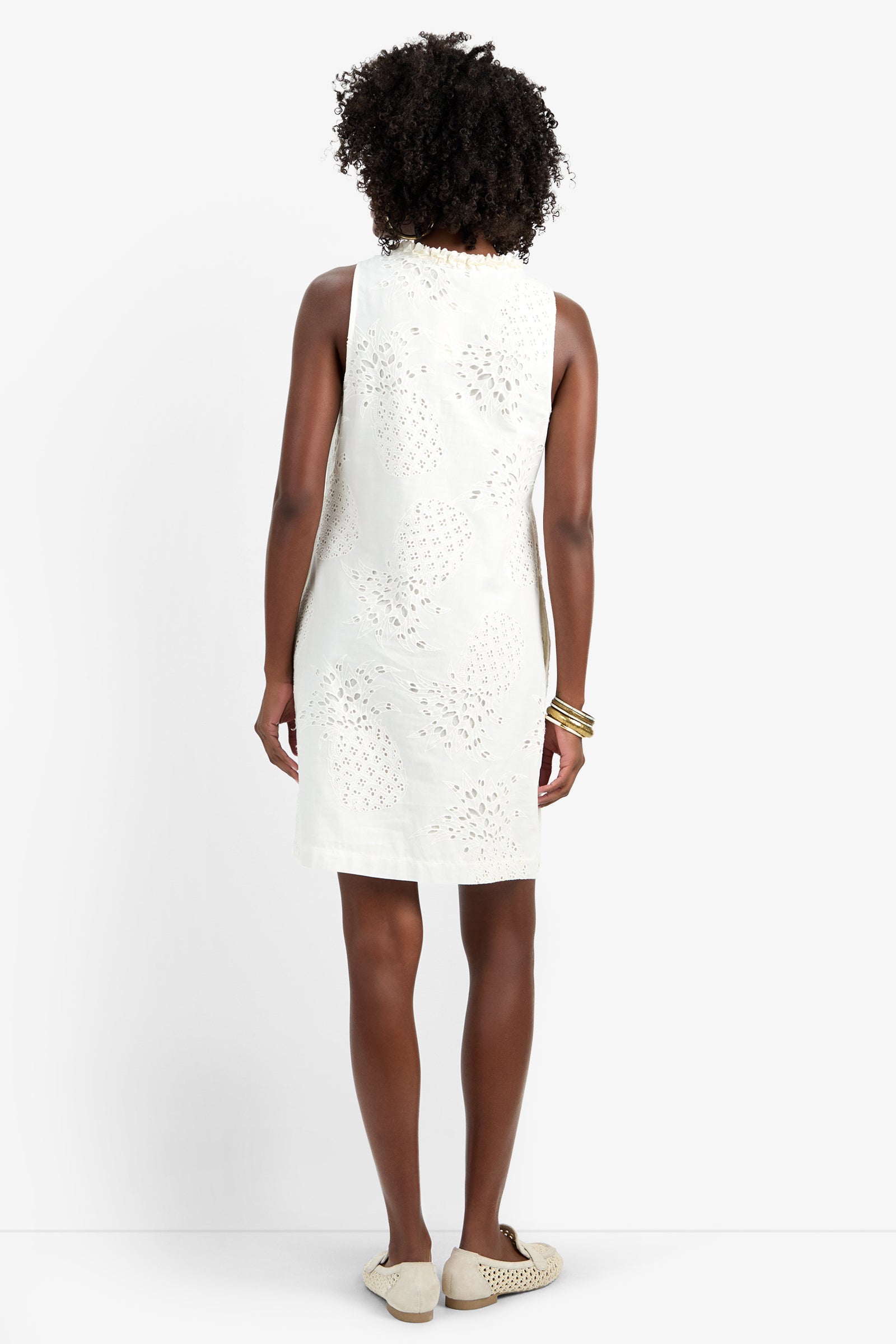 A woman with curly hair stands with her back to the camera, wearing the sleeveless, knee-length AMALA DRESS in white 100% cotton eyelet, paired with white slip-on shoes and gold bangles, set against a plain white background.