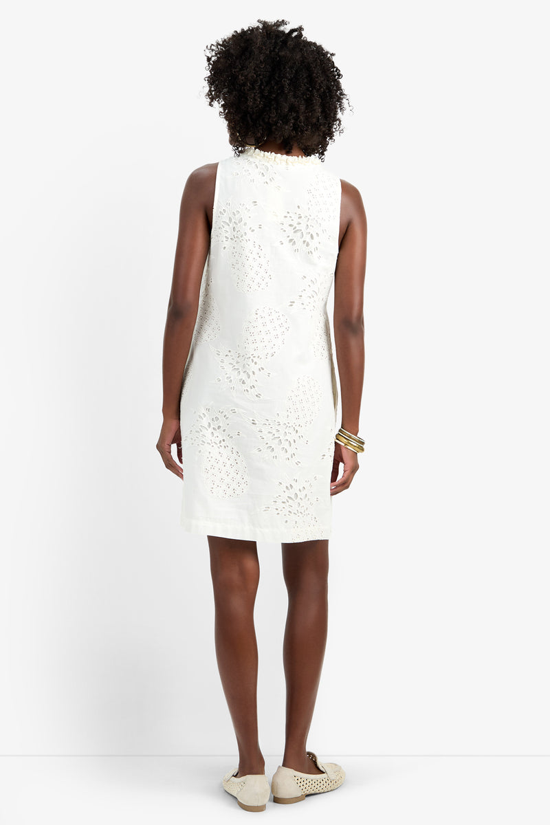 A woman with curly hair stands with her back to the camera, wearing the sleeveless, knee-length AMALA DRESS in white 100% cotton eyelet, paired with white slip-on shoes and gold bangles, set against a plain white background.