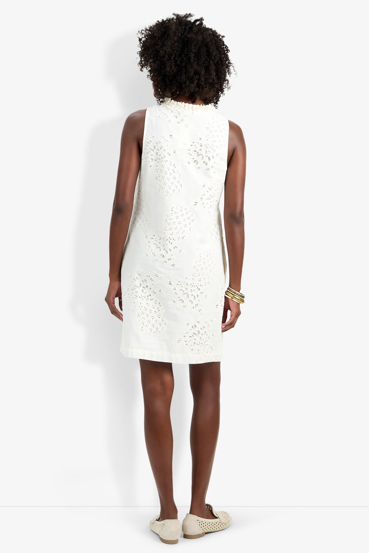A woman with curly hair stands facing away against a plain white background, wearing the white sleeveless knee-length Amala Dress, cream slip-on shoes, and several gold bangles on her left wrist.