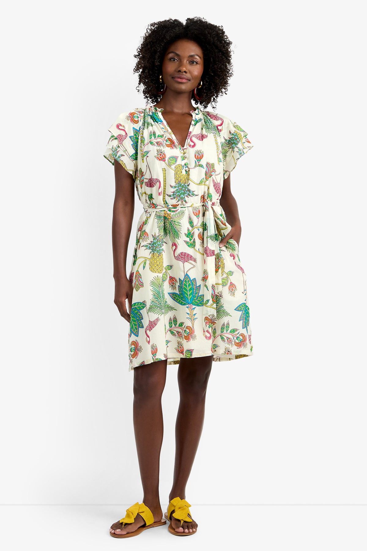 A woman stands against a white background wearing the ZANDY DRESS—a short, white 100% cotton dress with colorful tropical prints, short sleeves, a belted waist—and yellow sandals.