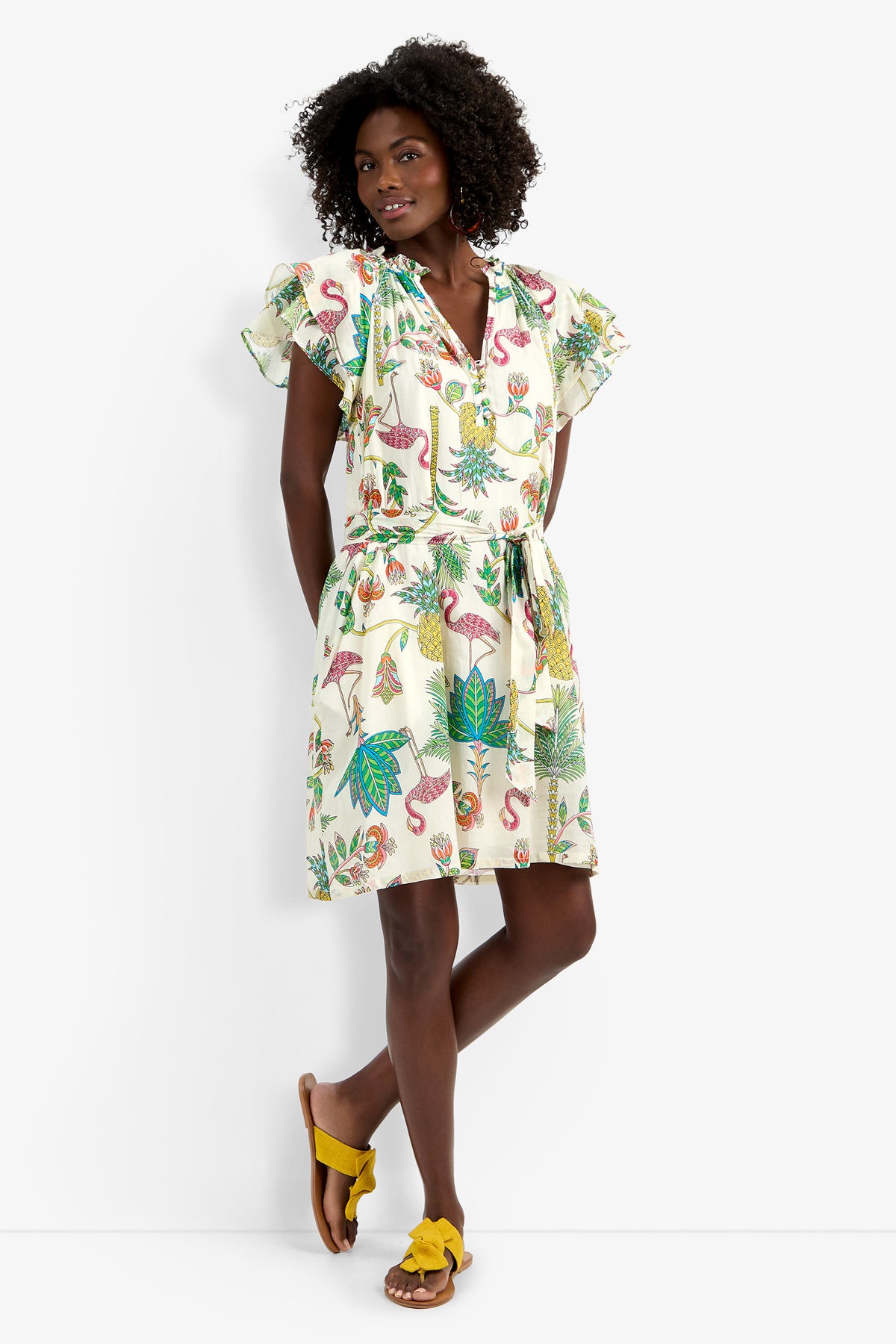 A woman with curly hair poses confidently against a white wall, wearing the Zandy Dress—white with colorful tropical prints, ruffled short sleeves, a belted waist—and yellow bow sandals.