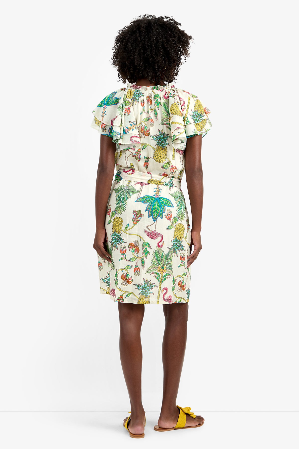 A person with curly hair stands facing away, wearing the ZANDY DRESS—a white 100% cotton dress with colorful tropical prints of leaves, pineapples, and flamingos. Ruffled short sleeves and yellow flat sandals complete the vibrant look.