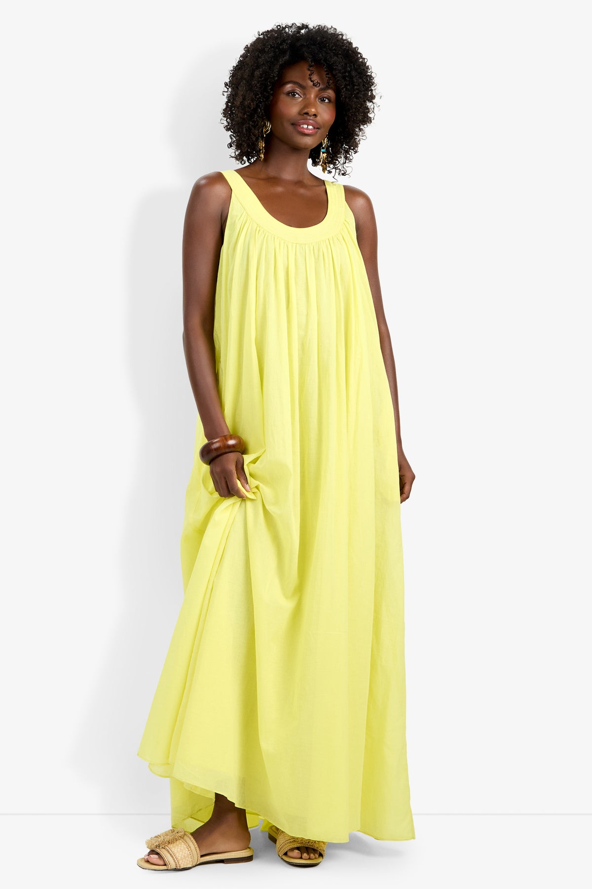 A woman with curly hair wears the Jianna Dress, a flowing sleeveless yellow maxi, paired with beige sandals. She stands against a plain white background, gently holding the dress and smiling softly.