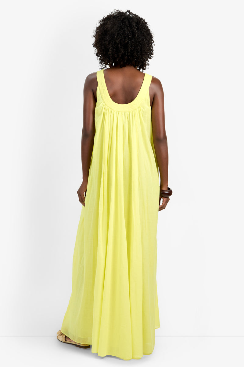 A woman with curly hair, facing away, wears the JIANNA DRESS—a sleeveless, long, loose-fitting yellow dress made of 100% cotton—with light-colored sandals against a white background.