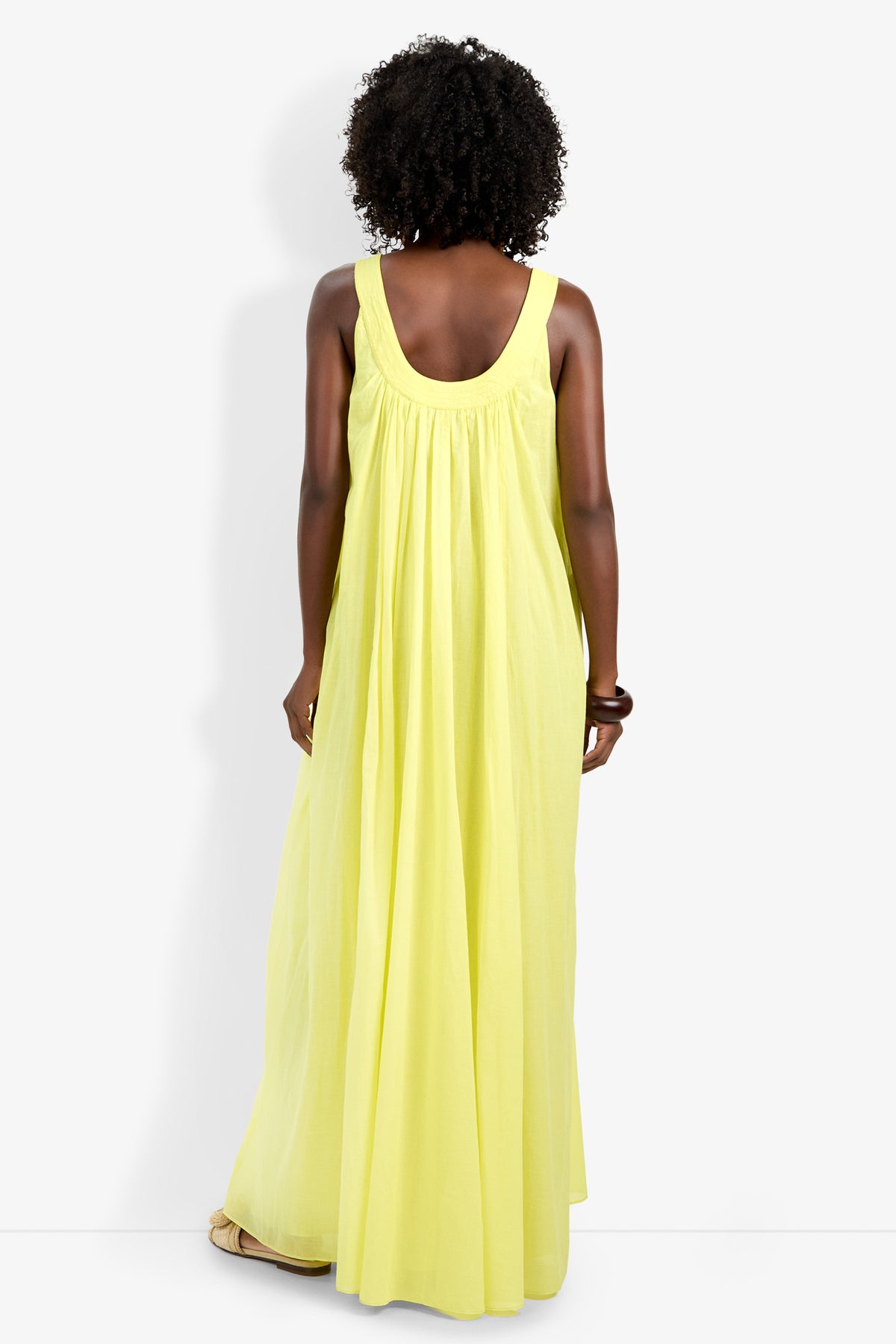 A woman with curly hair, seen from the back, wears the sleeveless yellow Jianna Dress and beige sandals, standing against a plain white background.