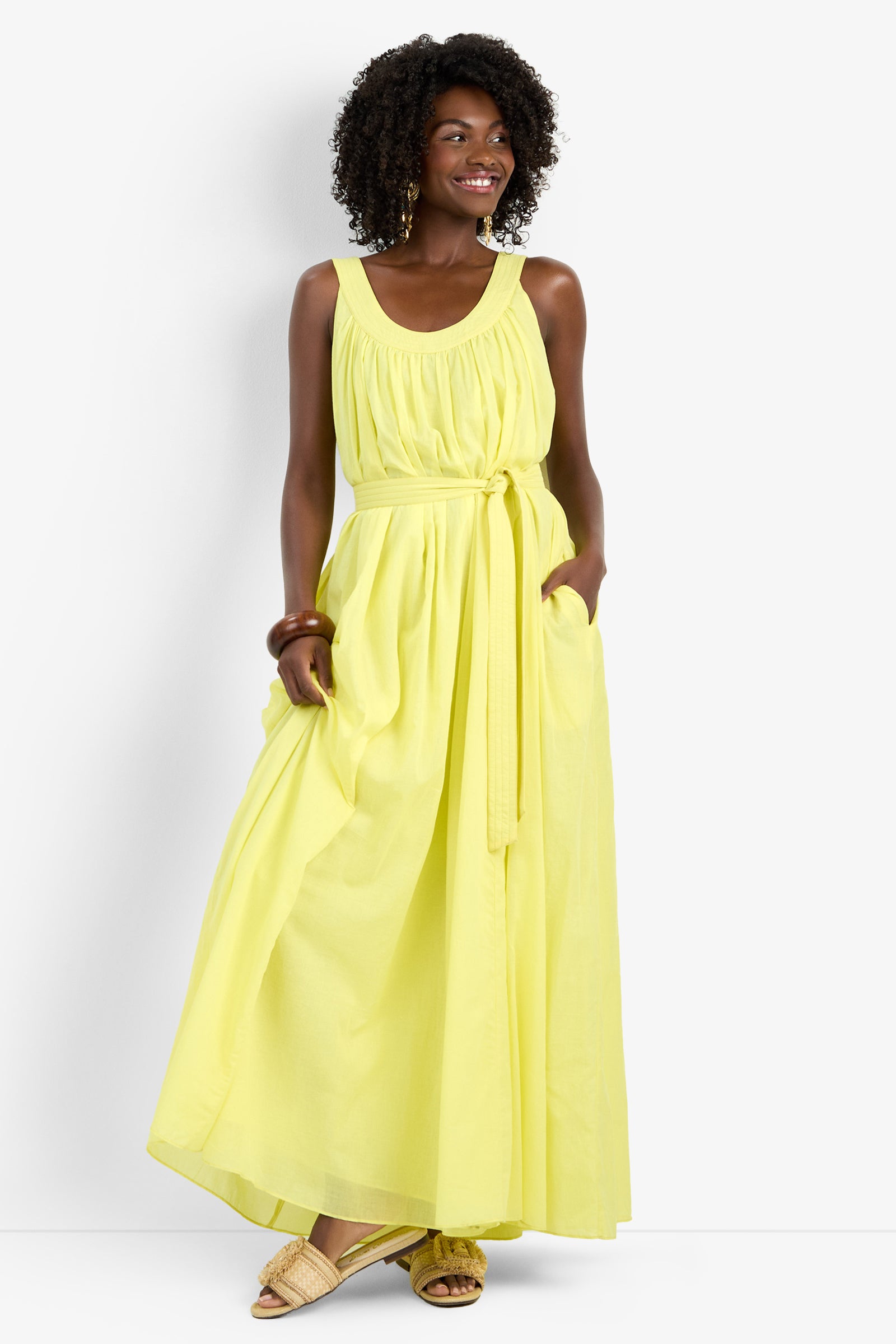 A woman smiles in the JIANNA DRESS, a sleeveless, floor-length light yellow 100% cotton dress with a gathered waist tie and pockets. She pairs it with light woven sandals and a bracelet.