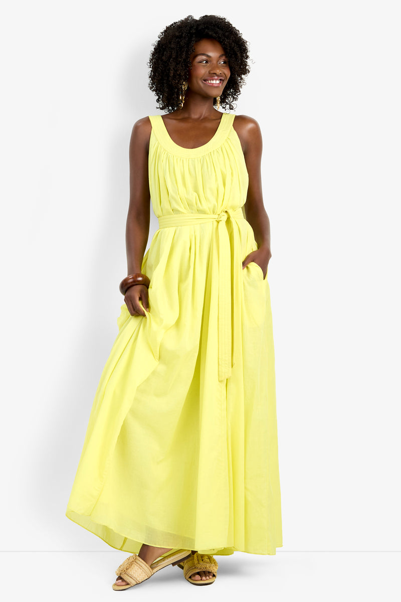 Woman smiling and looking to the side, wearing the sleeveless yellow Jianna Dress with a belted waist and side pockets; she has curly hair, tan flat sandals, and a brown bracelet.