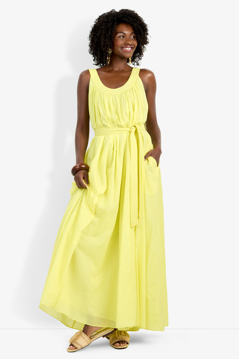 A woman models the Jianna Dress—a sleeveless, bright yellow maxi with a tied waist and pockets. She smiles, wearing gold earrings and sandals, with curly hair, against a plain white background.