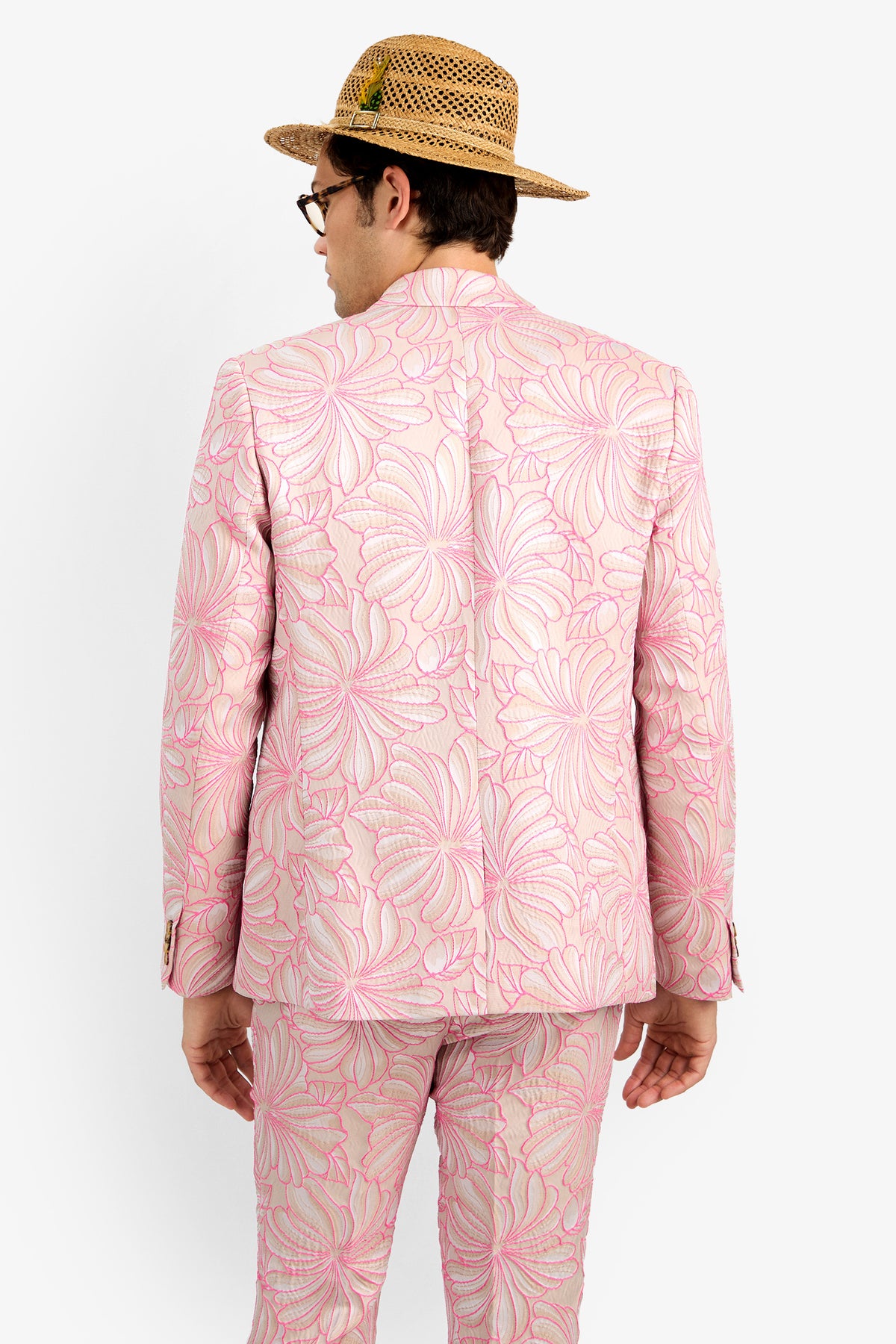 A person wearing the THURSTON BLAZER, featuring a bold floral pattern in light pink, and a straw hat stands facing away from the camera against a plain white background.
