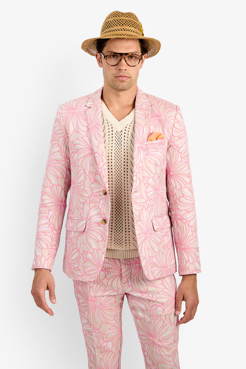 A man wearing the THURSTON BLAZER, a pink floral-patterned suit jacket, with a beige knit shirt, straw hat, and glasses stands against a plain white background, looking directly at the camera with a neutral expression.