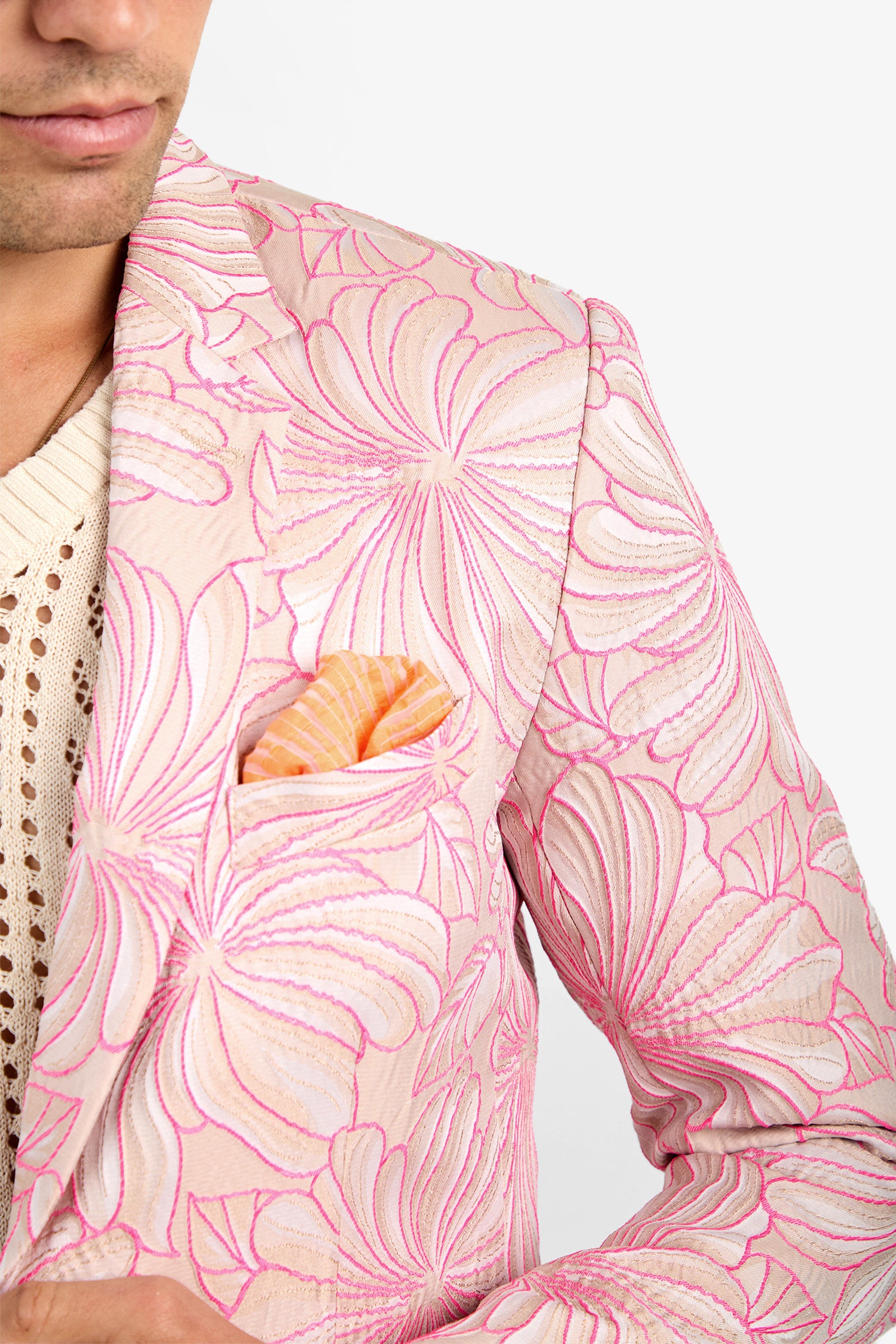 A person wearing the THURSTON BLAZER—a beige jacket with bold pink floral embroidery—over a cream knit top and an orange-striped pocket square. Only their lower face and upper body are shown.